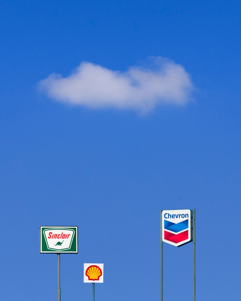 three filling station signs against a blue sky containing a single cloud