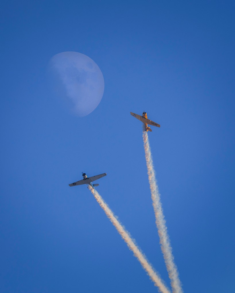 two aerobatic airplanes with a half-moon in the background